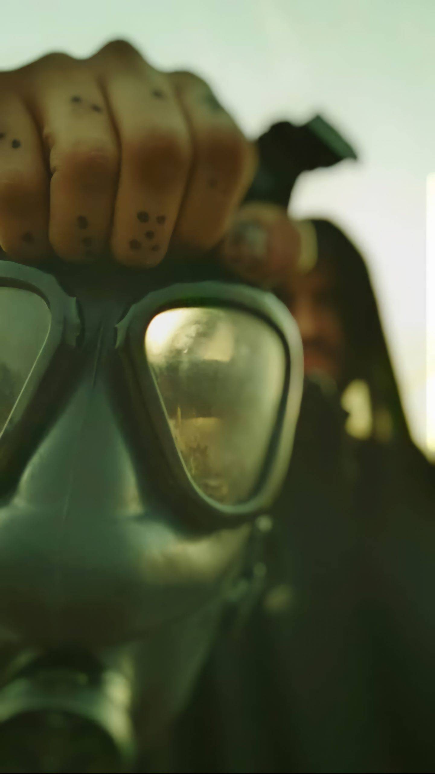 Close-up of a person in a gas mask holding the top of it with tattooed fingers, shot in moody green lighting.