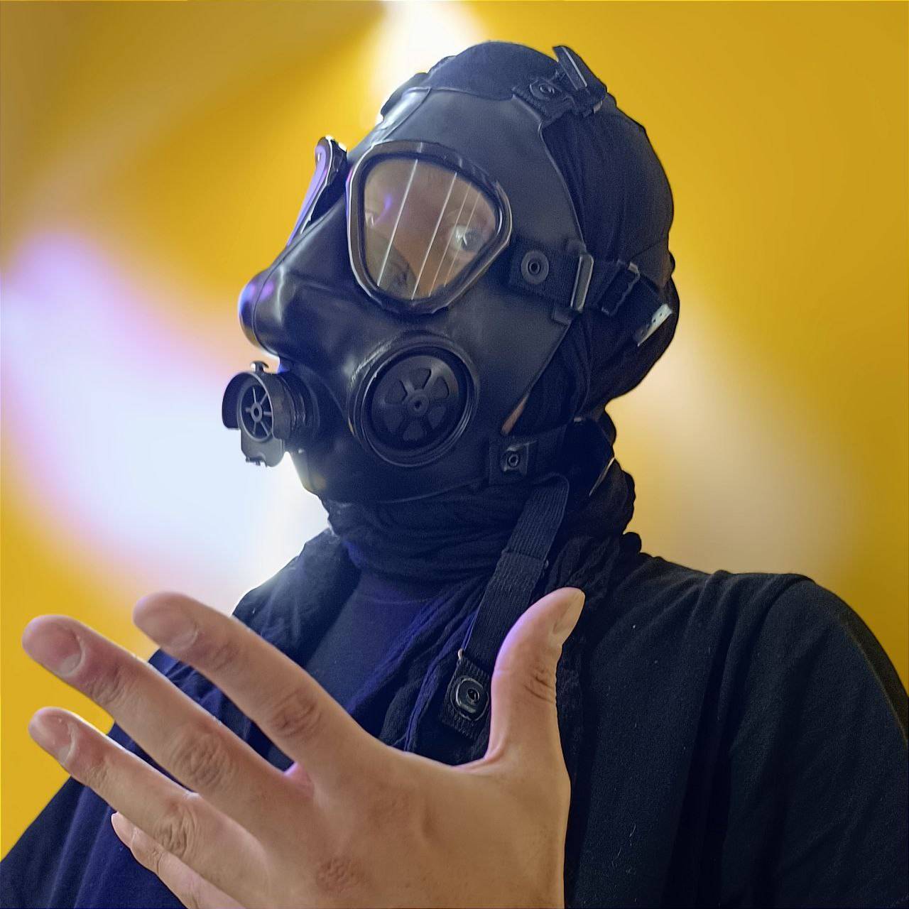 Wayloun band member in a gas mask and black clothing, gesturing with one hand against a vivid yellow background.